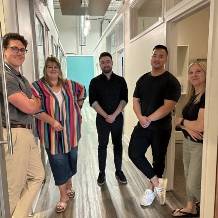 A group of five people is standing together in a hallway, smiling and posing casually.