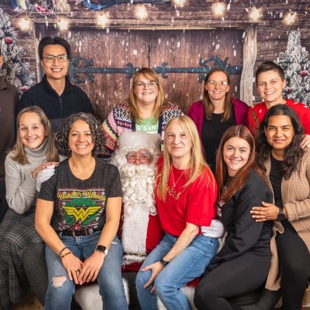 A festive group photo with eleven people posing in front of a holiday-themed backdrop with Christmas decorations. The group includes a mix of men and women, and Santa Claus is seated in the middle. Everyone is smiling and wearing holiday-themed attire, with a few people sitting and others standing around them.