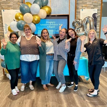 A group of six people is celebrating in front of a display with balloons and a "Happy Anniversary to Align!" message on a screen. They are all smiling and posing for the photo, with one person holding a large silver "20" balloon to mark the 20th anniversary.